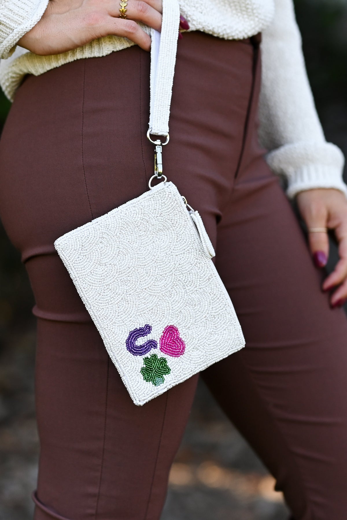 A person wearing brown pants and a cream sweater carries a white textured crossbody bag with a white strap. The bag features embroidered purple and pink letters and a green shamrock on the front, set against a blurred outdoor background.