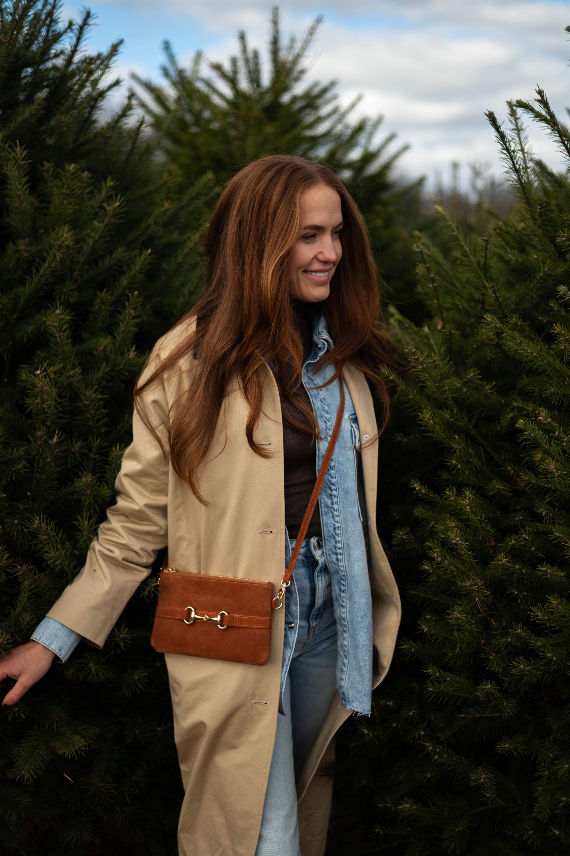 Woman standing among Christmas trees wearing a beige coat and brown handbag.